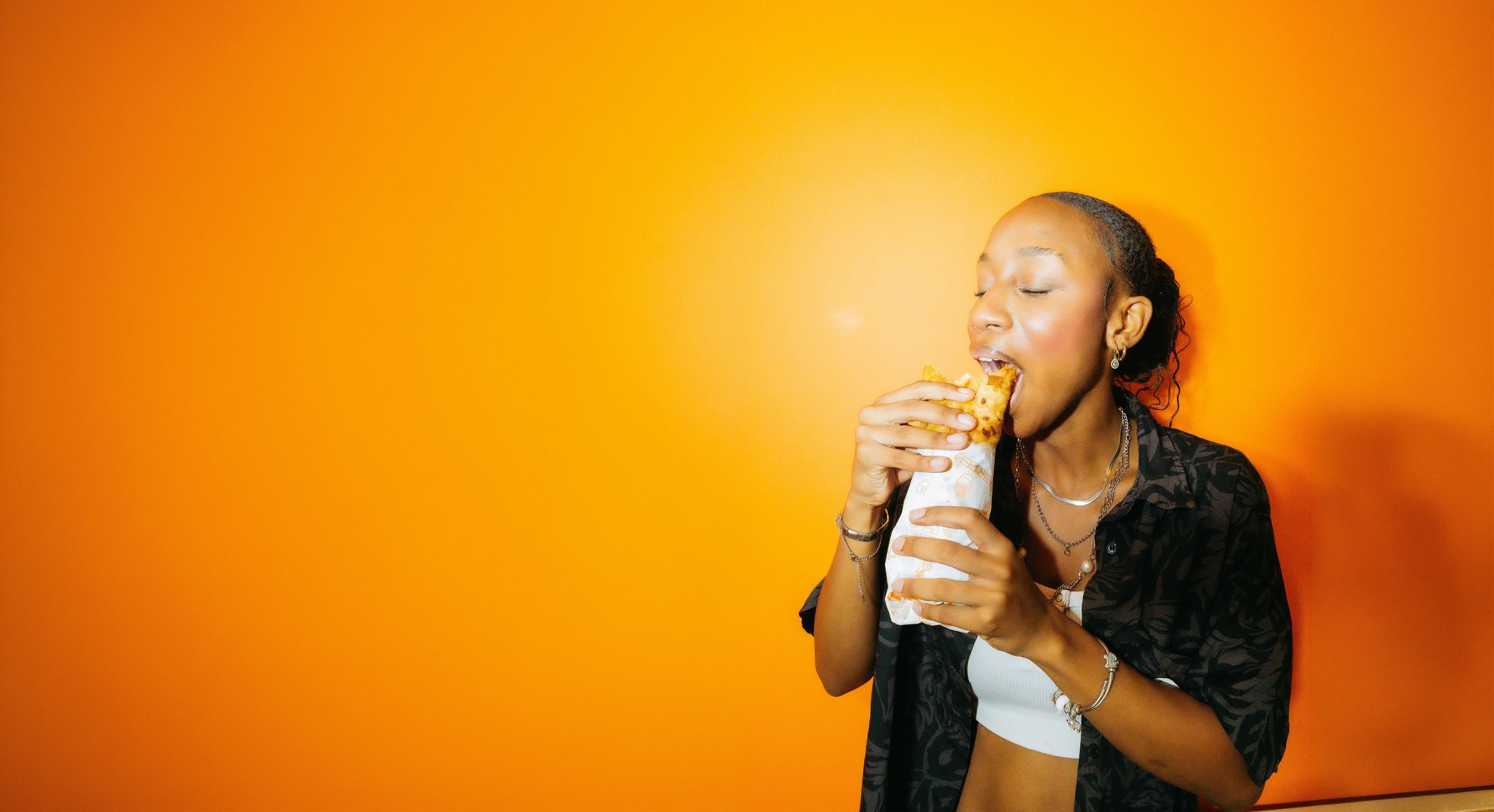 A woman taking a big bite of a wrap against an orange wall